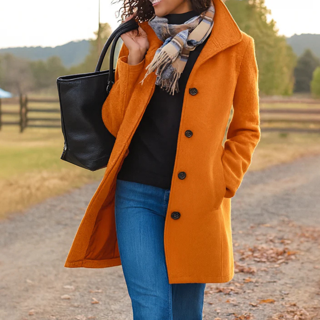 Orange version of the coat with a textured finish, worn with a black top, jeans and checked scarf on a country path. 7