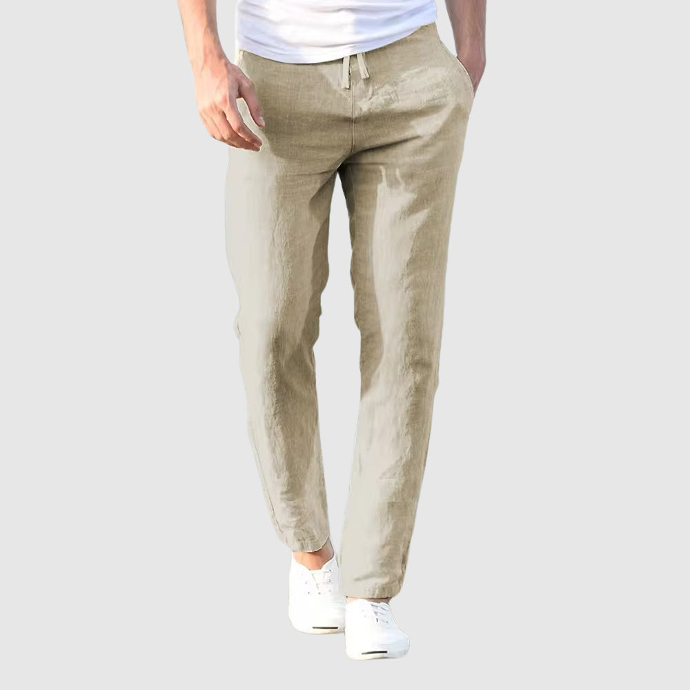 Front view of the trousers in pale stone beige on a light grey background, worn with a white T-shirt and white plimsolls. 2