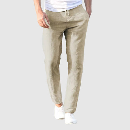 Front view of the trousers in pale stone beige on a light grey background, worn with a white T-shirt and white plimsolls. 2