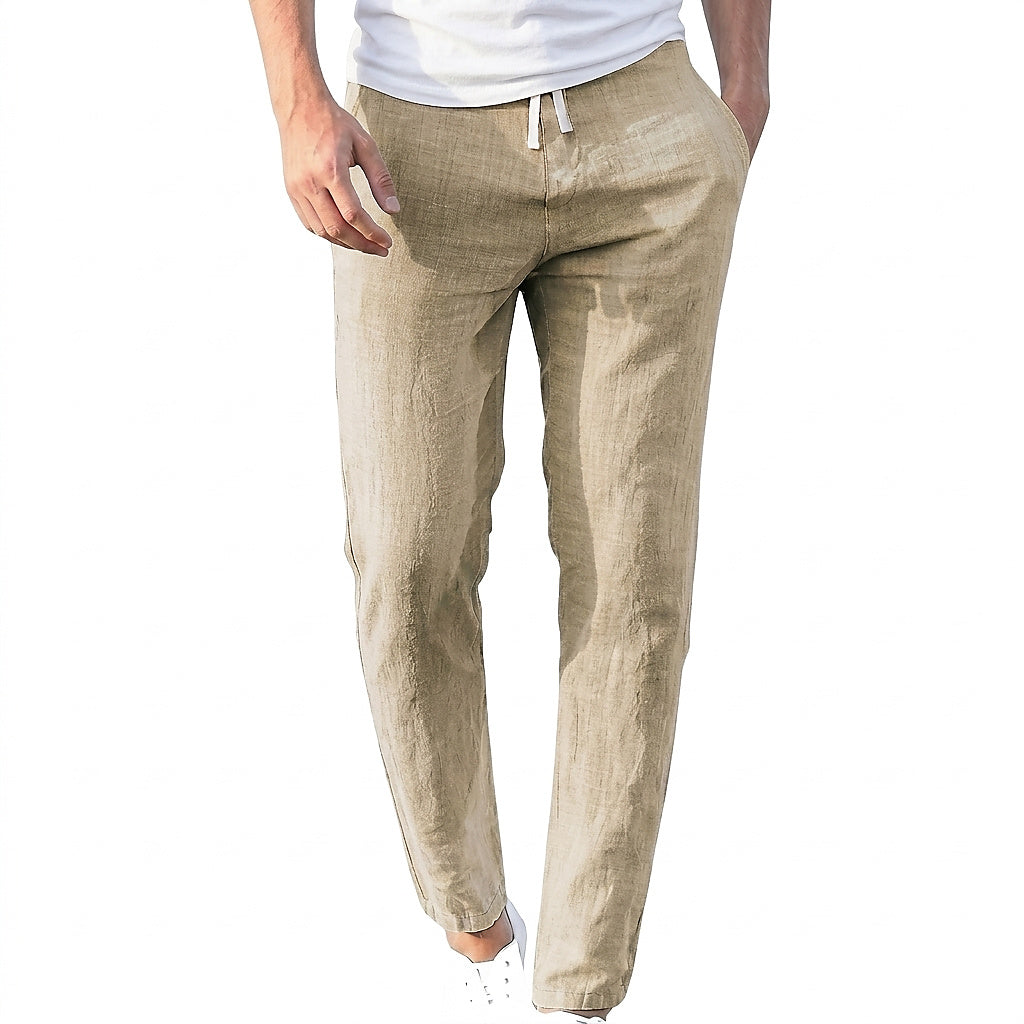 Model wearing sand-coloured, slightly creased linen trousers with a white drawstring, a white T-shirt and white trainers, one hand in a pocket. 1