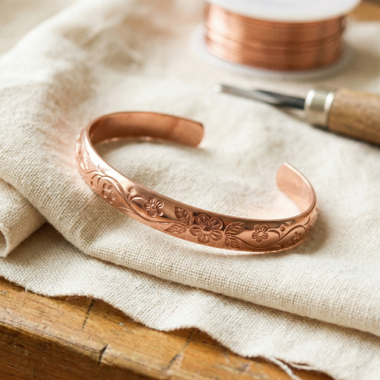 Open copper cuff with engraved flowers, leaves and curling vines, resting on linen beside a spool of wire 1