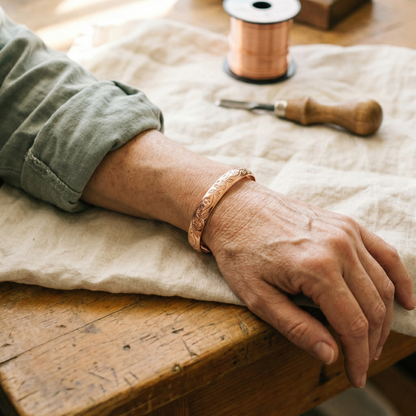 Copper cuff with engraved floral pattern worn on a wrist resting on a linen-covered wooden table 2
