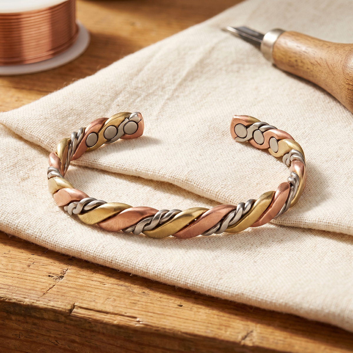 Open cuff of twisted rose copper, gold-brass and silver-tone strands with round magnets at both ends, resting on cream linen 1