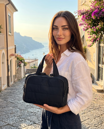 Smiling woman in a white linen shirt holds a black version of the quilted bag, with a sea view behind her. 8