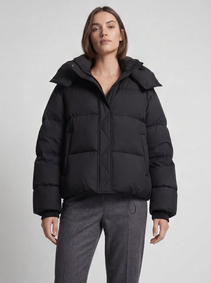 Close front view of a model in the black oversized puffer jacket, with grey trousers and the hem drawcords visible 6