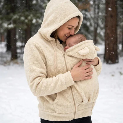 Woman in a cream sherpa hoodie with hood up kissing her baby's head in the carrier panel, in a snowy forest. 8