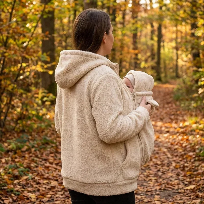 Rear three-quarter view of a woman in a cream sherpa hoodie on a woodland path, baby in the bear-eared carrier panel. 6