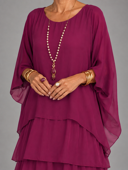 Upper body detail in magenta, showing the round neckline, flowing cape sleeves and layered hem, worn with pearl necklace. 24