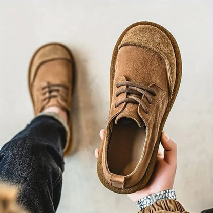 Hand holding one light tan suede-look lace-up shoe, with the other shoe worn on a foot behind 1