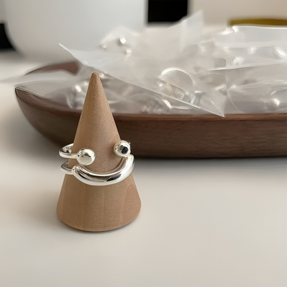 Silver smile ring displayed on a wooden cone stand, with a wooden dish of small bagged rings softly blurred behind. 7