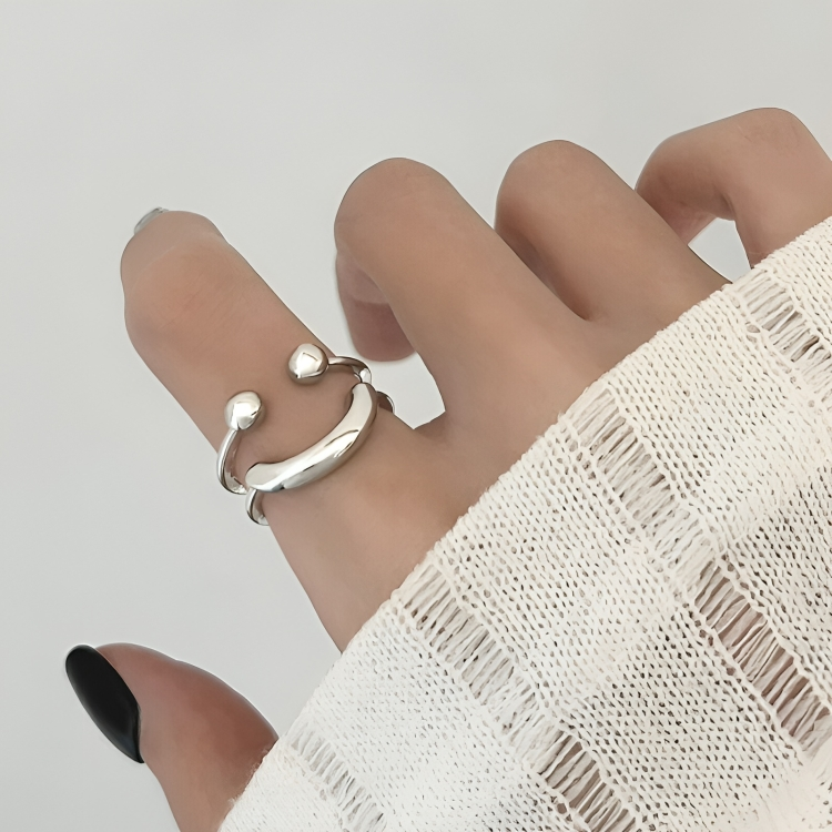 Close-up of the open silver ring on a finger, its two polished balls and curved bar seen from the side, beside a cream knit sleeve. 3
