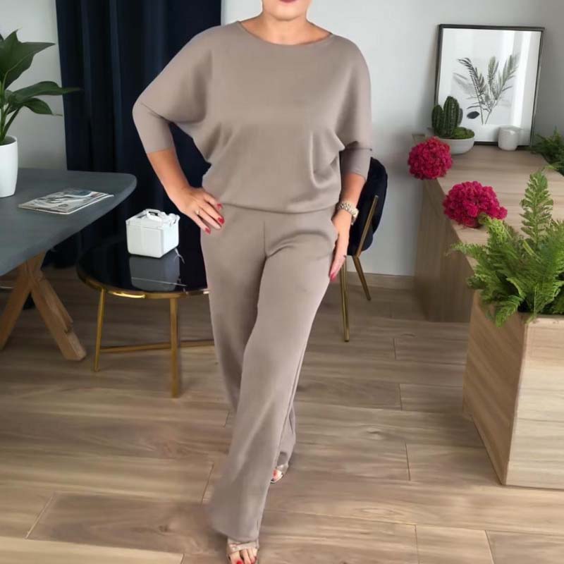 Woman in a taupe batwing-sleeve top and matching wide-leg trousers, hand on hip, standing in a living room. 8