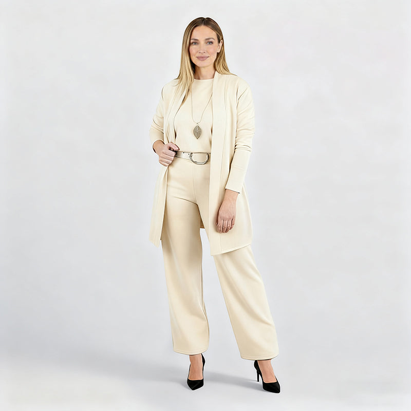 Full-length studio shot of a model in the cream set with belt, leaf pendant and black heels, hand holding the cardigan. 14