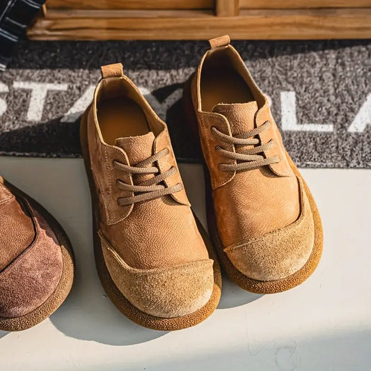 Tan lace-up shoes with suede toe caps and chunky soles, seen from above on a pale floor beside a doormat. 2