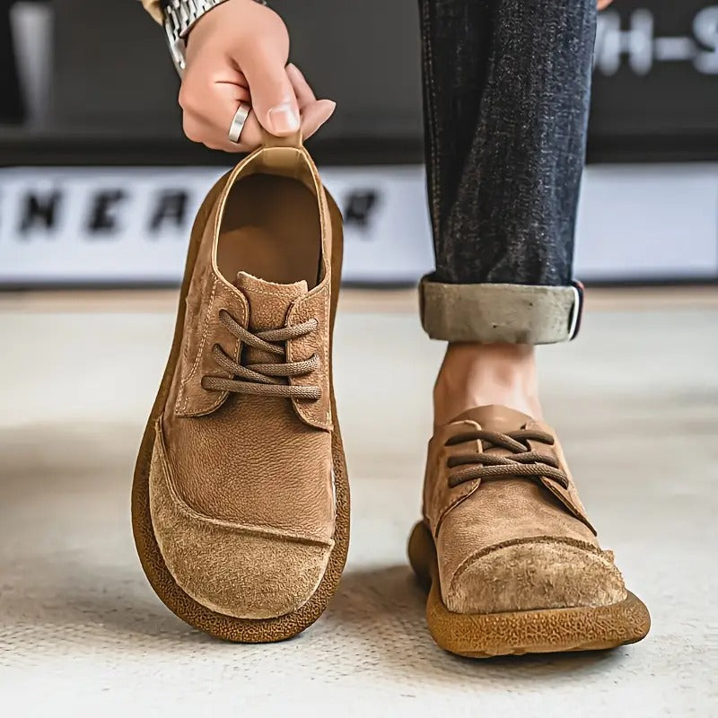 Man holding one tan suede lace-up shoe by its heel tab while wearing the other, with rolled-up dark jeans. 3
