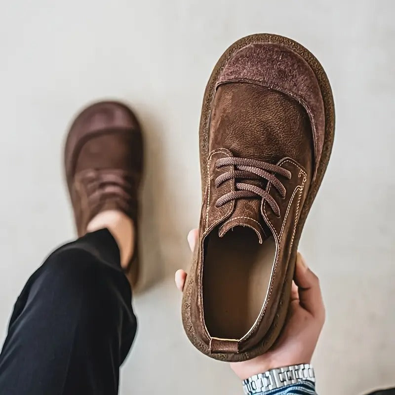 Dark brown suede shoe held in one hand showing its stitched side and lace-up front, other foot worn nearby. 7