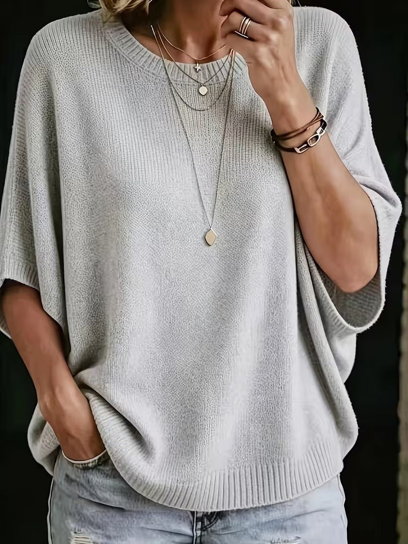 Close-up of the light grey knit batwing top worn with layered necklaces and jeans, one hand raised to the chin. 1