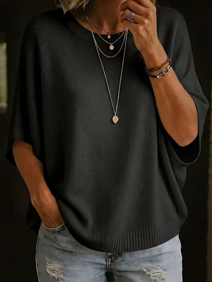 Close front view of the black knit batwing top on a model wearing layered gold necklaces and bracelets, one hand at chin. 26