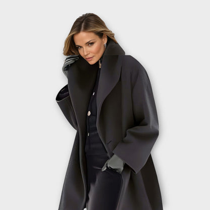 Model in the coat in black with a broad shawl collar, worn open over dark clothing and grey gloves. 3
