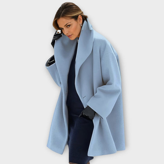Woman wearing the oversized wide-lapel coat in pale blue over a navy dress, with black leather gloves. 1