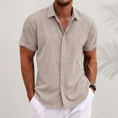 Taupe textured short-sleeved shirt worn by a model with hands in white trousers, against a pale wall with palm shadow. 2