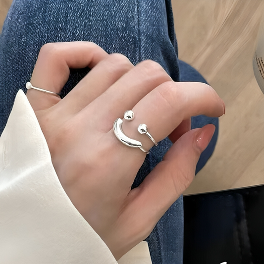 Silver open ring with two ball ends and a curved bar forming a smile, worn on a forefinger beside a thin pearl ring. 1