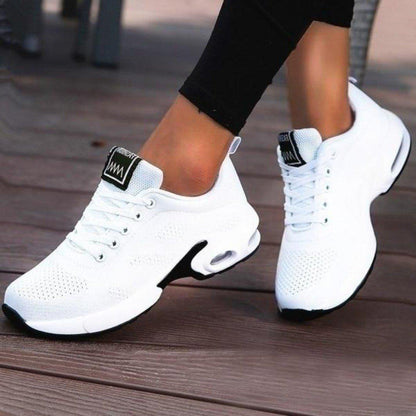 Plain white mesh trainers with a black-edged air-cushion sole and white laces, worn with black leggings on wooden decking. 6