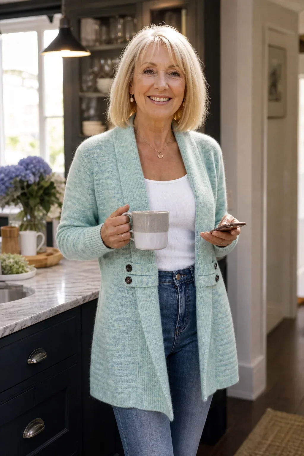 Older woman smiling in a light mint-green cardigan, holding a mug and phone in a kitchen 4