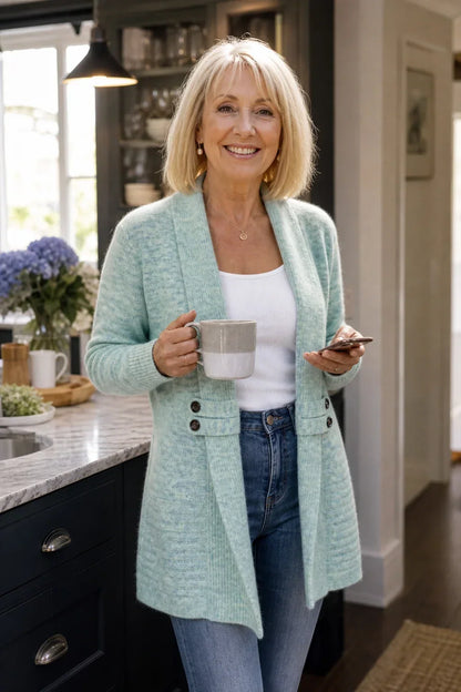 Older woman smiling in a light mint-green cardigan, holding a mug and phone in a kitchen 4
