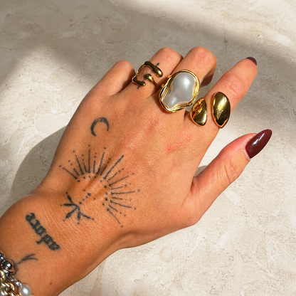 Tattooed hand on a pale stone surface wearing the gold-toned open ring alongside a large pearl-style ring and a wavy gold ring. 4