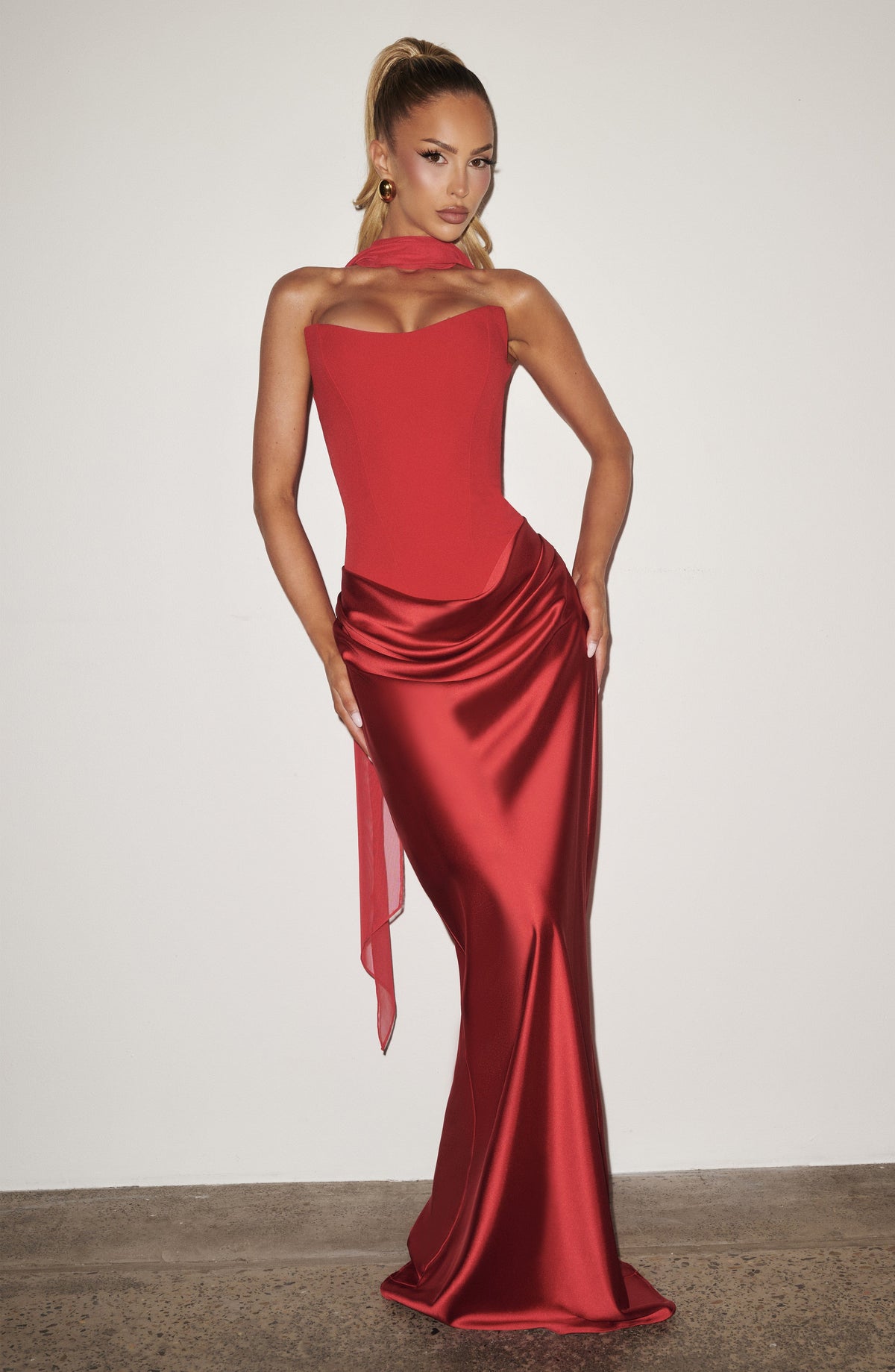 Blonde model in the red corset dress and scarf posing against a plain white wall 14