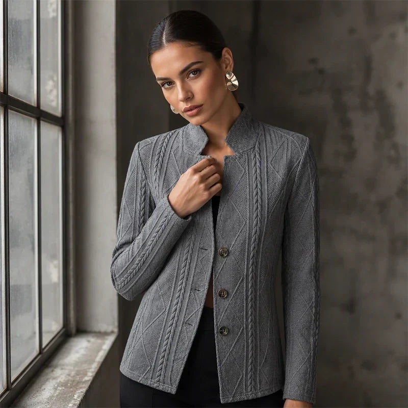 Grey blazer with cable pattern, model touching the collar beside a window in a moody interior 10