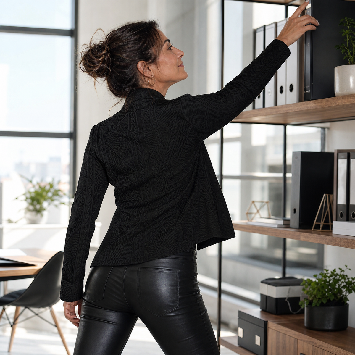 Back view of the black textured blazer, worn with leather-look trousers while reaching for an office shelf 13