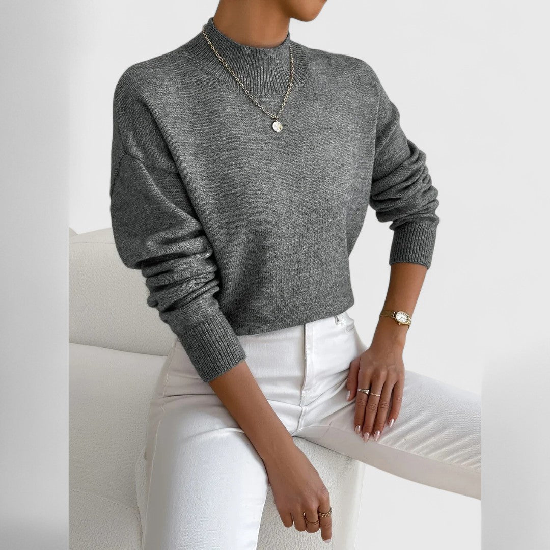 Heathered grey jumper worn by a model seated on a white chair, with a ribbed high neck, white jeans and a gold pendant. 12