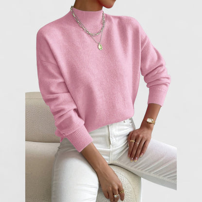 Soft pink jumper on a seated model in white jeans, showing the ribbed funnel neck and cuffs with silver necklaces. 5