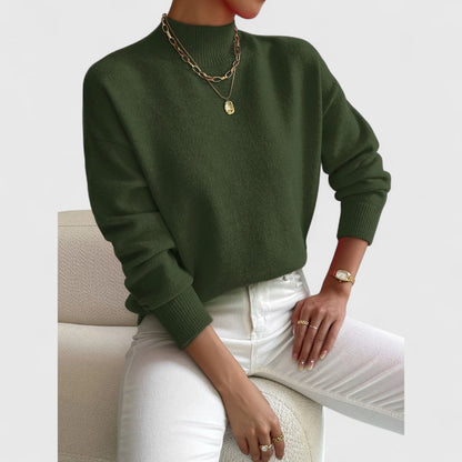 Dark olive green jumper worn by a seated model with white jeans, showing the ribbed high neck and gold necklaces. 10