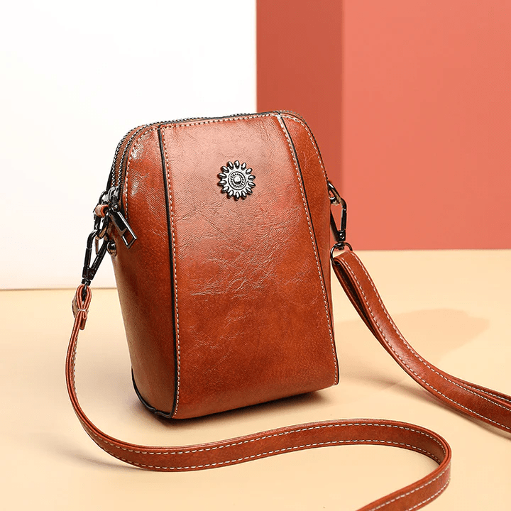Tan leather crossbody bag with a daisy stud, seen at an angle with strap trailing 1