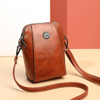 Tan leather crossbody bag with a daisy stud, seen at an angle with strap trailing 1