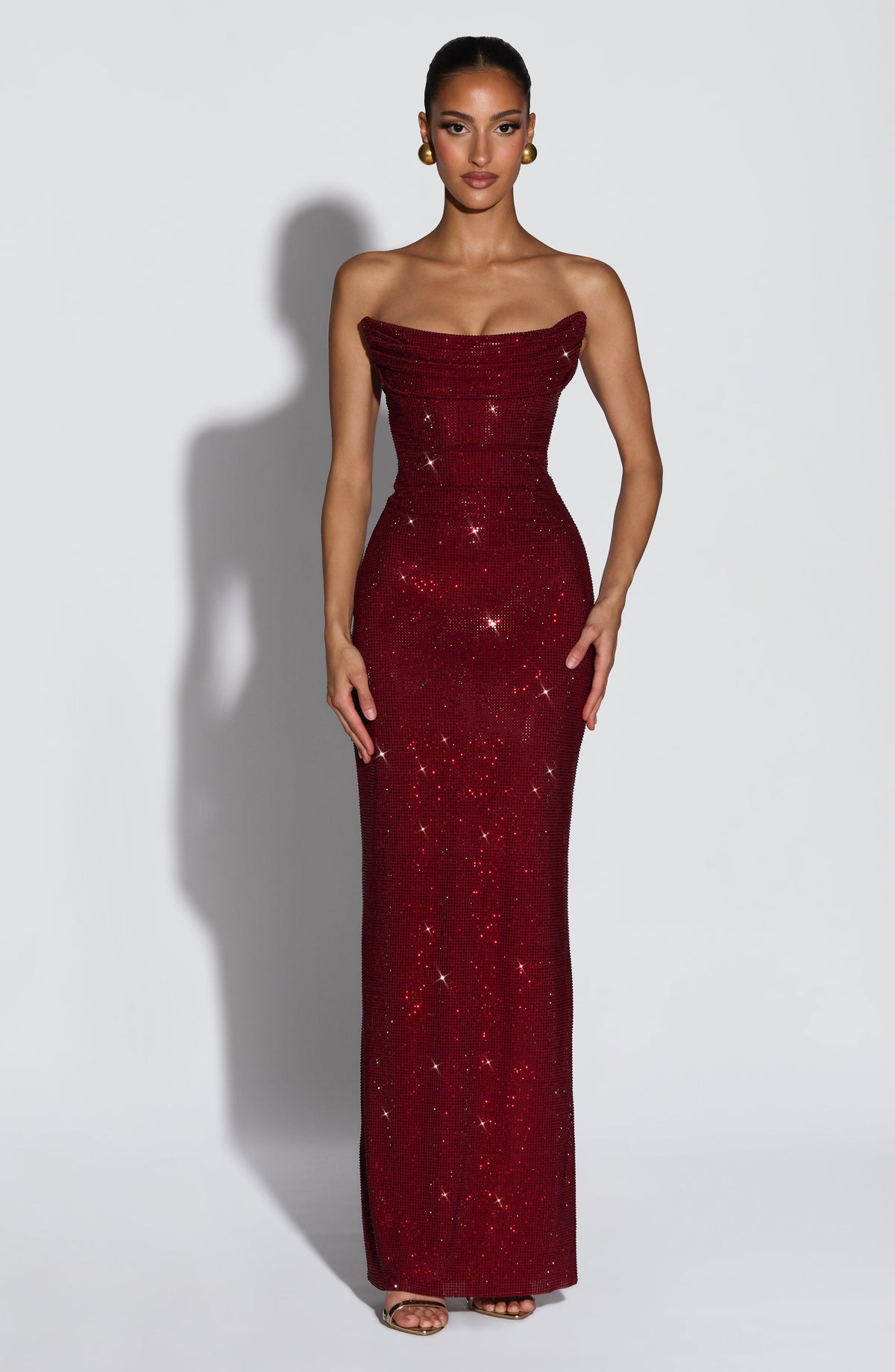 Model faces front in the burgundy rhinestone maxi dress, strapless draped neckline, gold earrings and heels 1