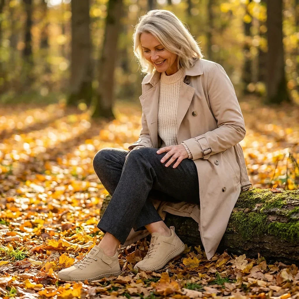 Smiling woman in beige coat sitting on a mossy log in autumn leaves, wearing beige trainers and looking down. 13