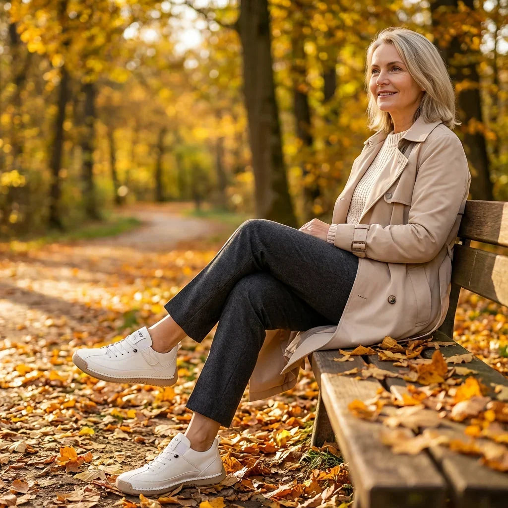 Woman in beige trench coat sitting on a park bench among autumn leaves, wearing white trainers and crossing her legs. 12