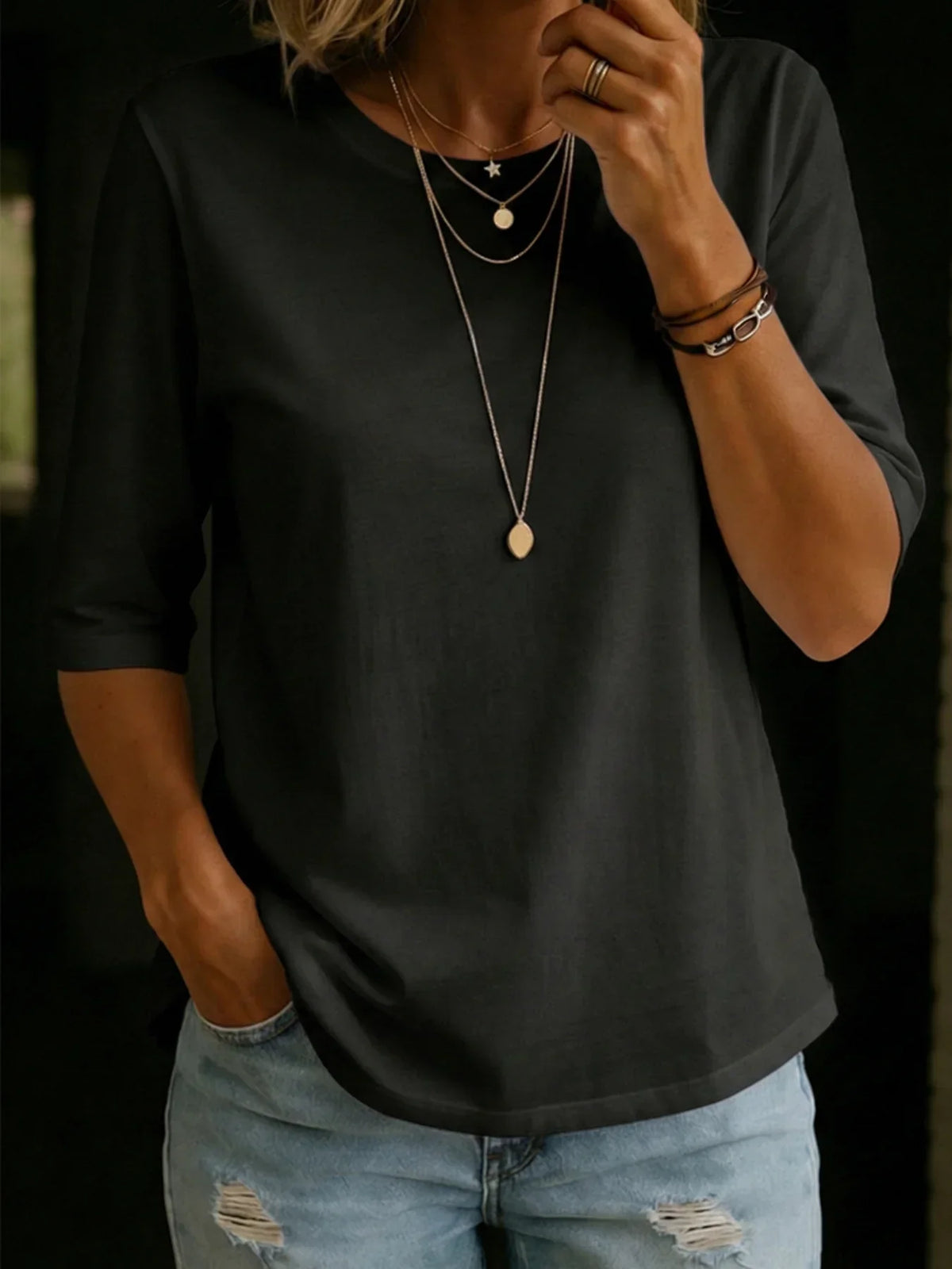 Woman wearing the black half-sleeve crew-neck tee with layered gold necklaces, stacked bracelets and ripped light-wash jeans. 4