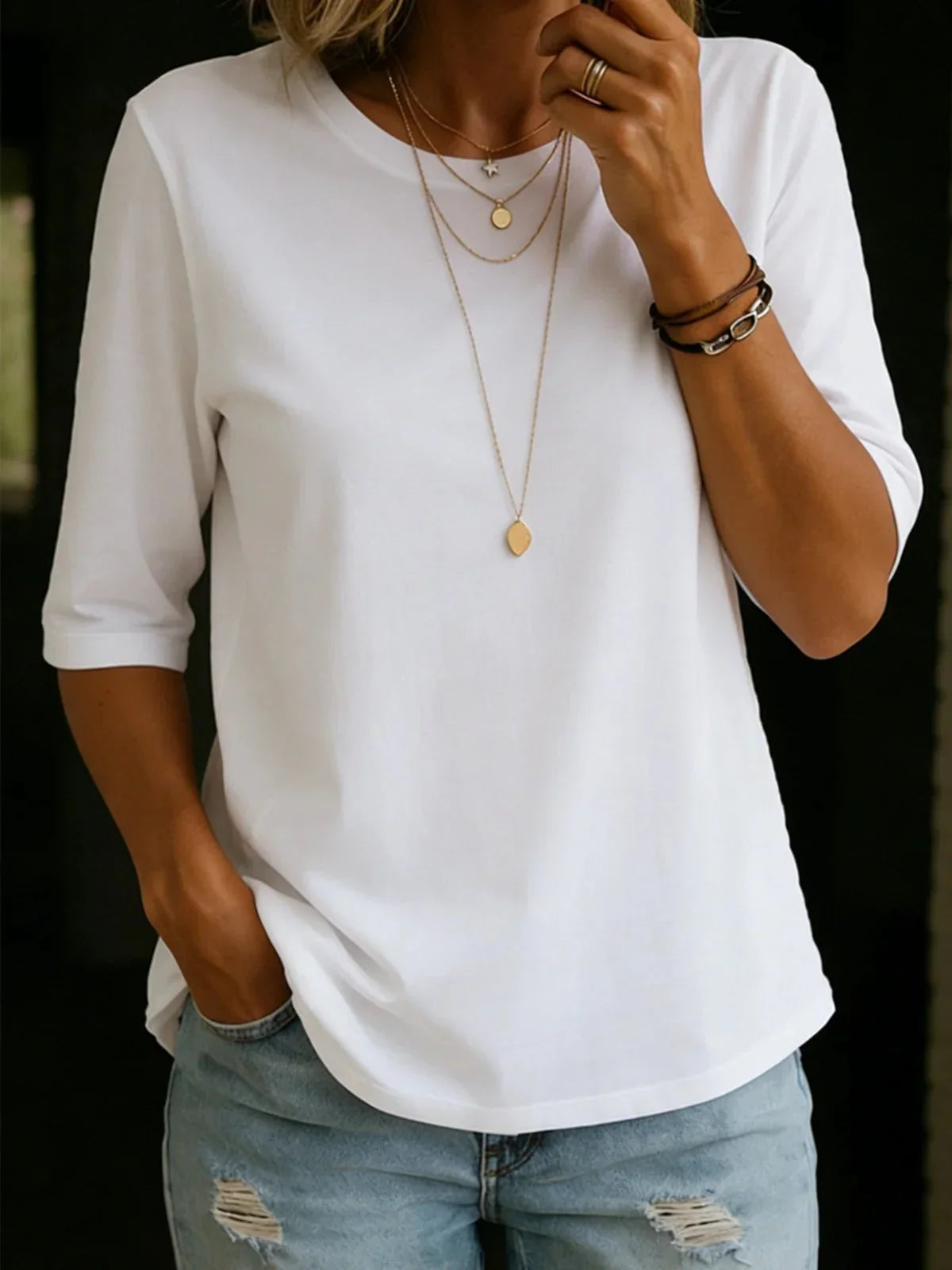 Woman wearing the white half-sleeve crew-neck tee with layered gold necklaces and ripped light-wash jeans, one hand in her pocket. 1