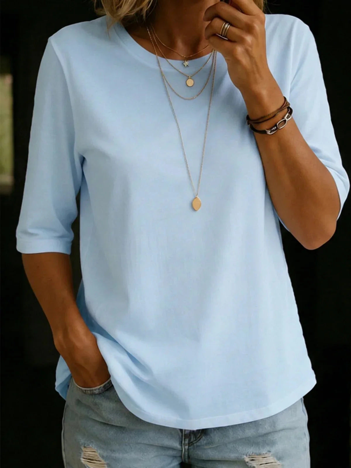 Woman wearing the pale blue half-sleeve crew-neck tee with layered gold necklaces, bracelets and ripped light-wash jeans. 2