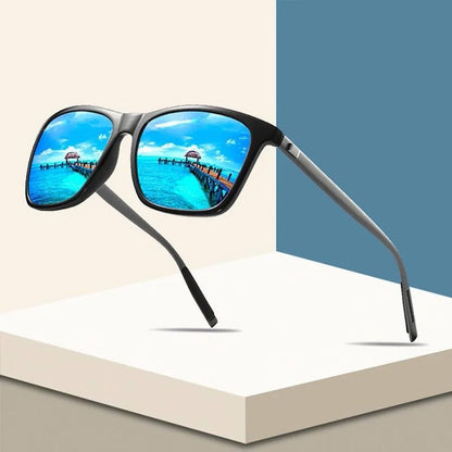 Black square-framed sunglasses with blue mirrored lenses reflecting a tropical pier, displayed on pale geometric blocks 1