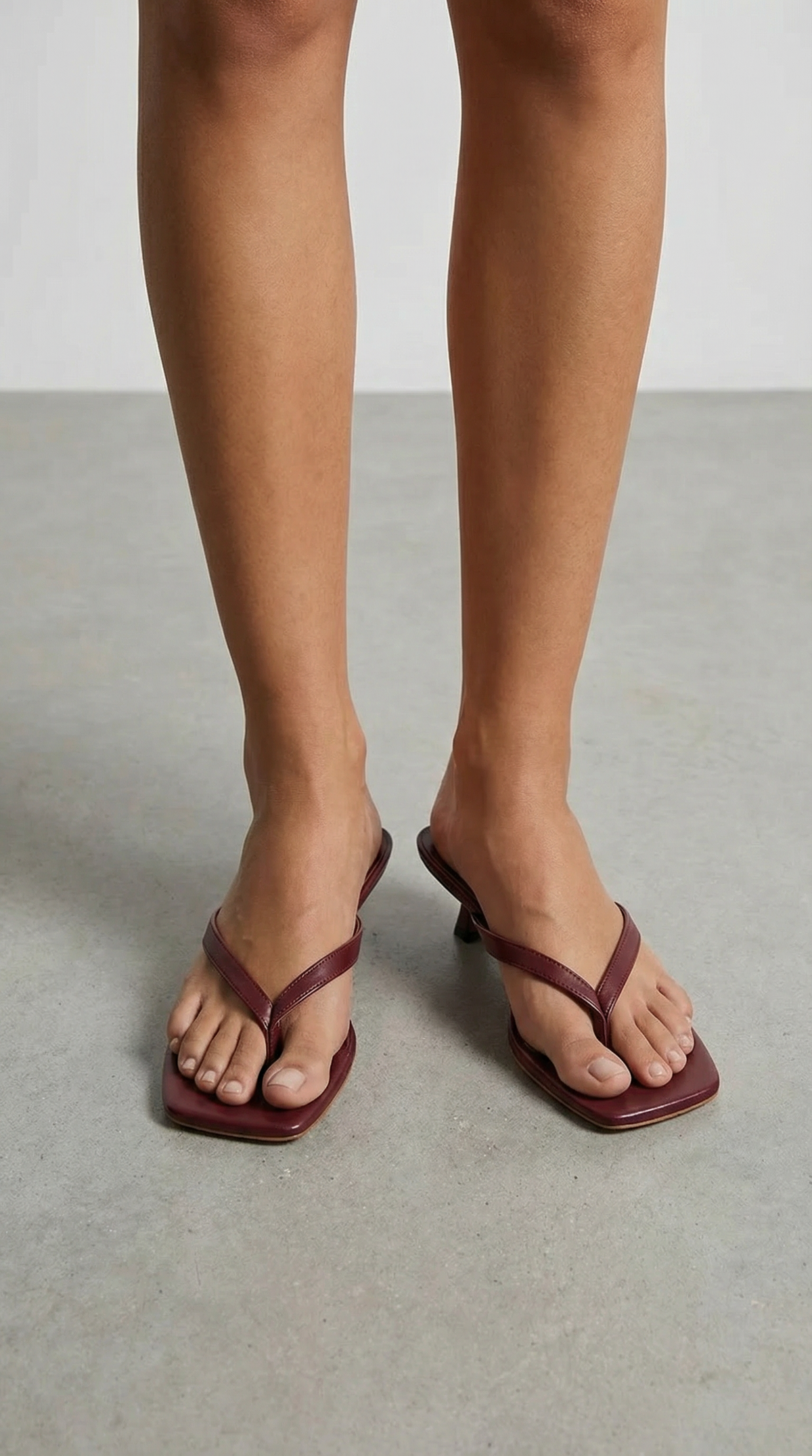Front view of a model's feet in burgundy kitten-heel thong sandals, standing on a grey concrete floor. 14