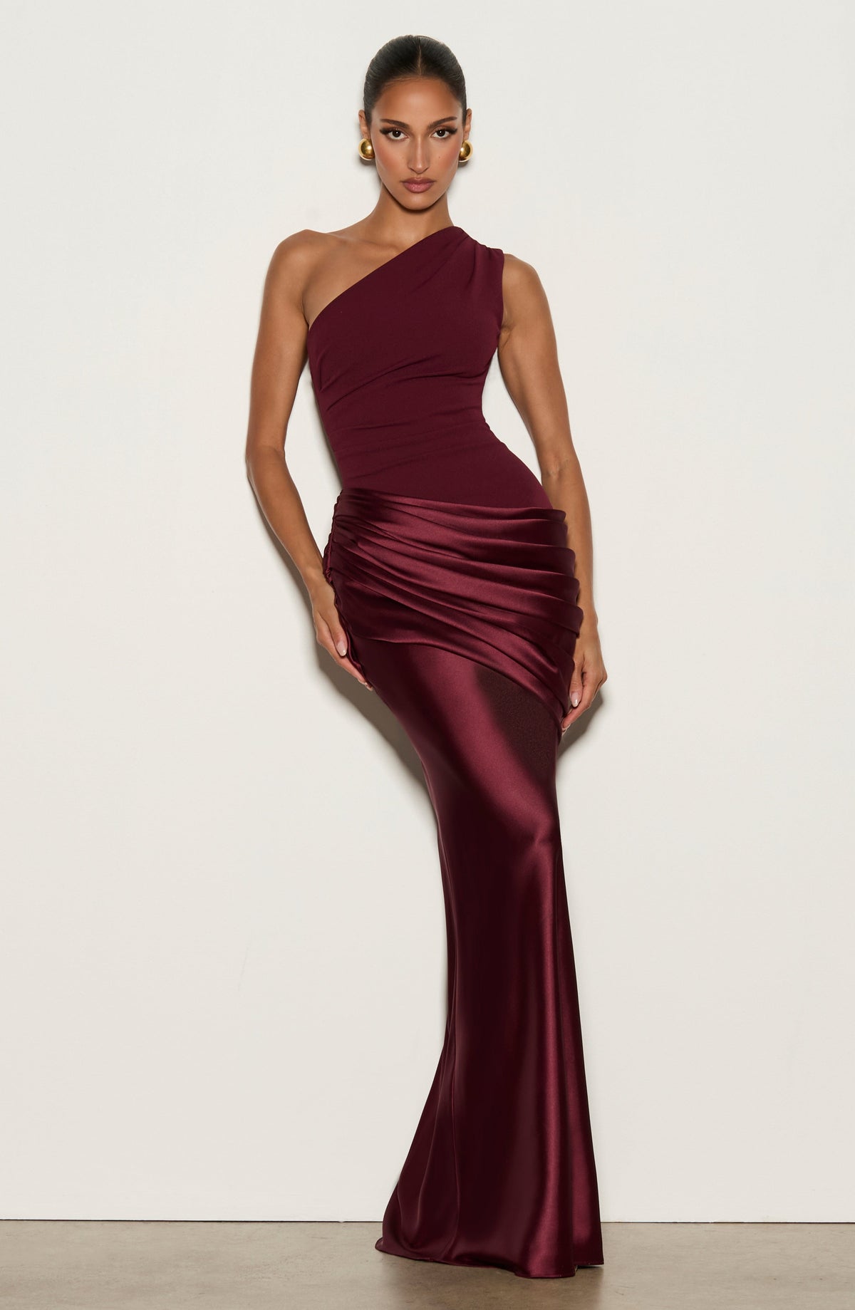 Model leaning against a white wall in the burgundy gown, with gold earrings 16