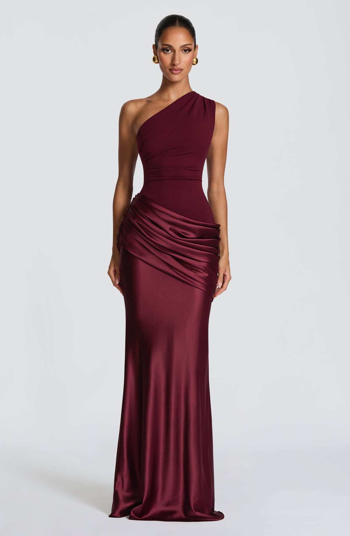 Burgundy one-shoulder maxi dress against a white wall, model leaning slightly with hands at her hips. 36
