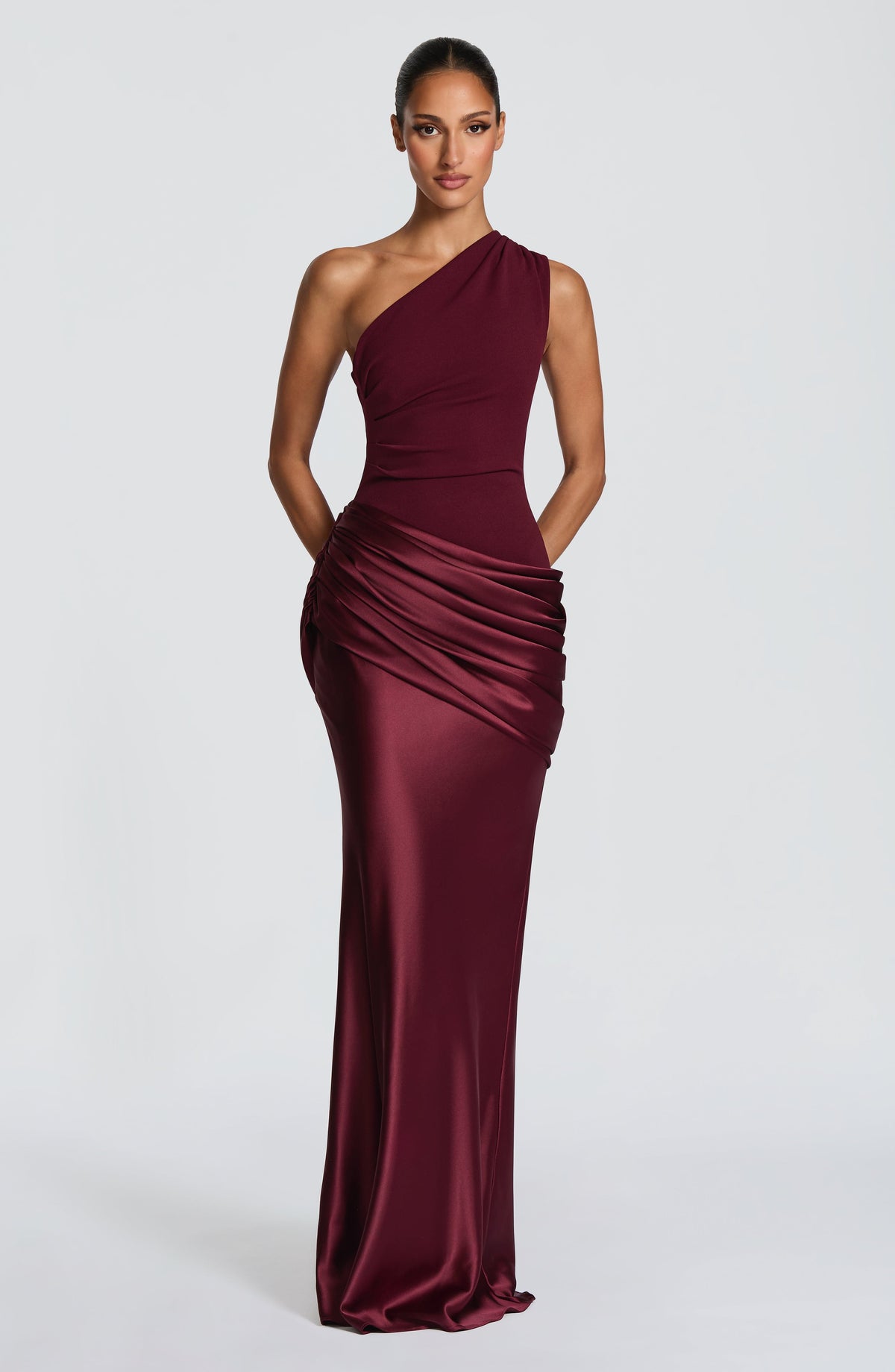 Burgundy one-shoulder dress against a plain wall, one arm raised to her head and the other behind her back. 38