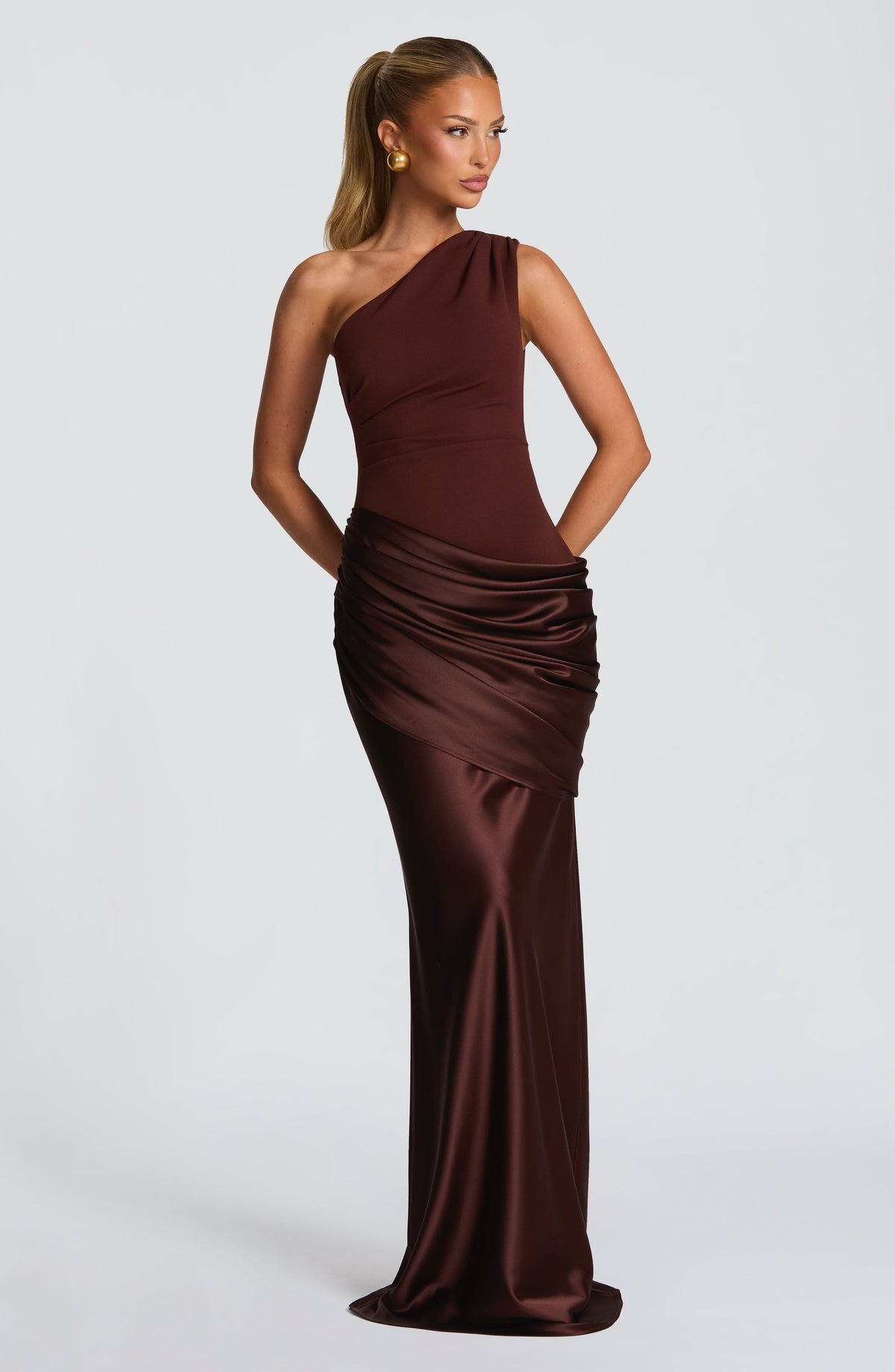 Model in the dark chocolate brown one-shoulder satin maxi dress with ruched hip drape, hands behind her back. 7